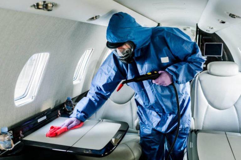 A cleaning person in blue protective suit wiping down surfaces aboard an airplane before flight.